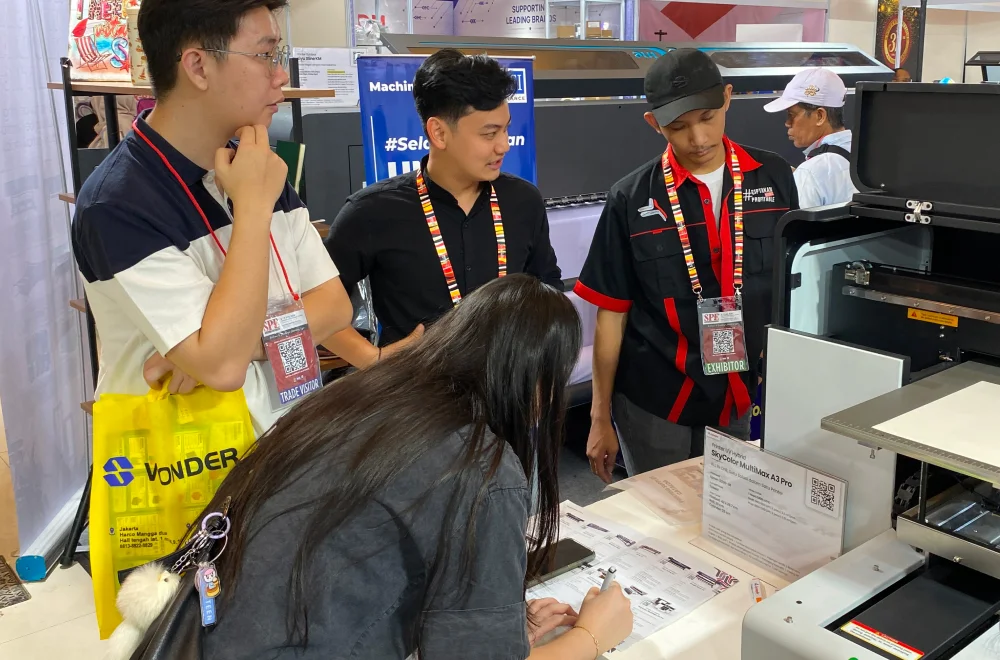 Pengunjung Berkonsultasi Dengan Tim Laysander Di Surabaya Printing Expo 2026 Sambil Melihat Demonstrasi Printer Uv Untuk Menemukan Solusi Cetak Yang Sesuai Kebutuhan Bisnis.